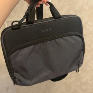 Svart och grå datorväska från Targus - Praktisk datorväska från Targus i svart och grå polyester. Väskan har ett stort huvudfack med dragkedja, ytterficka framtill och justerbar axelrem. Perfekt för att bära laptop och tillbehör. Snygg och stilren design med handtag och ID-ficka på baksidan.