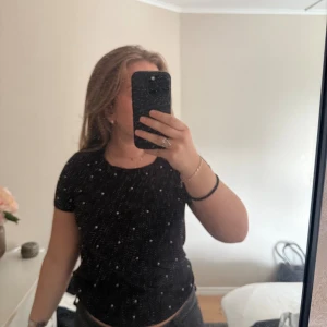 Svart croppad prickig topp - Svart croppad topp med korta ärmar och silvriga pärlor. Toppens material känns mjukt och stretchigt, perfekt för en chill och trendig look. Rund halsringning och figurnära passform som framhäver siluetten. 