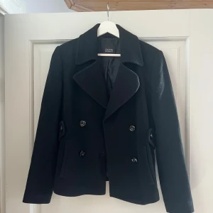 Svart skepparkavaj från Steffen Schraut - Svart skepparkavaj med dubbelknäppning och bred krage från Steffen Schraut. Jackan har snygga knappar fram och bak, samt fickor på sidorna. Tillverkad i ullblandning, perfekt för kyliga dagar. Klassisk och stilren modell med lång ärm.