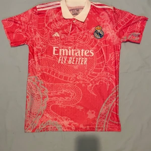 Real Madrid x Adidas rosa fotbollströja - Säljer en rosa Real Madrid fotbollströja från Adidas med vit krage och knappar. Tröjan har ett stort drakmönster över hela tyget, broderat klubbmärke och vita detaljer på axlarna. Materialet är lätt och andas, perfekt för träning eller match.