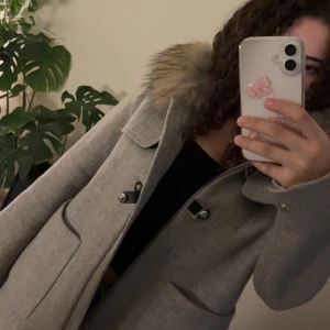 Ljusgrå kappa med fuskpäls - Snygg ljusgrå kappa med stor fuskpäls på kragen och coola detaljer med knappar och dragkedja. Jackan har stora fickor framtill och en rak, klassisk passform. Perfekt för dig som vill ha en stilren och trendig look under kallare dagar.
