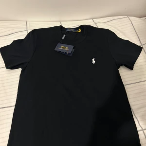 Svart t-shirt från Polo Ralph Lauren - Svart t-shirt från Polo Ralph Lauren med klassisk passform och rund hals. Ikoniska vita broderade loggan på bröstet. Tillverkad i mjuk bomull för en skön känsla. Perfekt basic-plagg med stilren look.