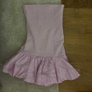 Axelbandslös rosa volangklänning - Supersöt axelbandslös kort klänning i ljusrosa med volang nertill. Klänningen har en figurnära passform upptill och en fluffig, rynkad kjol som ger en lekfull vibe. Perfekt på sommaren!!