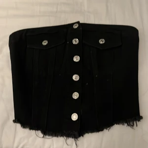 Svart jeanskorsett med silverknappar - Cool svart korsett i jeansmaterial med silverfärgade knappar framtill och rå fransig kant nedtill. Toppen har två dekorativa fickor med lock och knappdetaljer. Sitter snyggt och har en croppad passform som ger en edgy vibe.