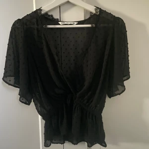 Svart omlottblus från Zara - Säljer en svart, transparent omlottblus från Zara med korta ärmar och små prickar i tyget. Blusen har spetsdetaljer vid halsringningen och resår i midjan för en snygg passform. Älskar denna men vuxit ur…