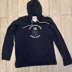 Marinblå hoodie från Björn Borg - Snygg marinblå hoodie från Björn Borg med vit text och tryck 'Stockholm Open' på bröstet. Tröjan har huva med vit snörning och känguruficka framtill. Perfekt för dig som gillar sportig och avslappnad stil. Skriv för fler bilder!!