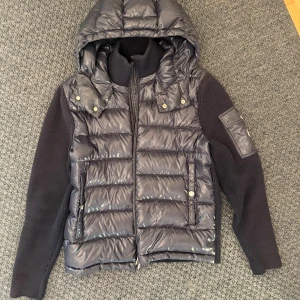 Moncler tricot cardigan - Snygg navy cardigan från Moncler med stickade ärmar och huva. Jackan är i nyskick o har quiltad front i glansigt material, dragkedja, fickor med tryckknappar och en ikonisk Moncler-logga på ärmen. Kvitto medföljer 