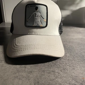 Vit och svart CR7 keps El Bicho - Snygg vit och svart truckerkeps med broderad patch framtill som visar en siluett av Cristiano Ronaldo och texten 'EL BICHO'. Baksidan är i svart mesh med CR7-märke. Perfekt för fotbollsfans som vill sticka ut.