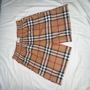 Burberry rutiga shorts med snörning - (Står Xl men riktiga sizet passar M-L)Snygga shorts från Burberry i klassiskt beige, svart, vit och röd rutig design. De har elastisk midja med svart snörning för perfekt passform. Tillverkade i mjuk bomull, perfekta för sommardagar. Ikonisk look som sticker ut.