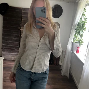 Randig vit skjorta med knappar - Snygg vit skjorta med tunna vertikala ränder i svart. Skjortan har lång ärm, klassisk krage och knäppning framtill. Passformen är normal och materialet känns lätt och skönt, perfekt till jeans eller kjol.