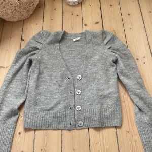 Grå kofta från Stories med knappar - Superfin grå kofta från Stories i storlek S. Koftan har v-ringning, ribbade muddar och fyra stora knappar framtill. Långärmad och i mjukt stickat material, perfekt för lager-på-lager under kyliga dagar.