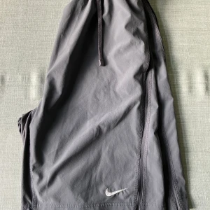 Grå träningsbyxor från Nike - Grå träningsbyxor från Nike med elastisk midja och dragsko. Byxorna är tillverkade i ett lätt och stretchigt material av polyester och elastan. Swoosh-logga broderad framtill. Perfekta för sport eller chill.