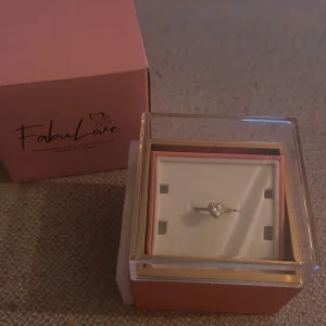 Hjärtformad silverring från FabuLove - Superfin guldfärgad ring från FabuLove med hjärtformad glittrande sten och små stenar runt om. Ringen har en tunn band och levereras i en söt ask med rosdetalj. Perfekt för dig som älskar romantiska smycken med extra bling.