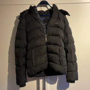 Svart pufferjacka från GANT - Svart pufferjacka från GANT med huva och dragkedja framtill. Jackan har två stora fickor och GANT-logga på bröstet samt längs dragkedjan. Perfekt för kalla dagar och riktigt snygg streetstil. Hör av dig vid minsta fundering 👍✅