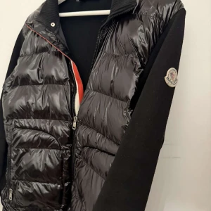 Svart pufferjacka från Moncler - Snygg svart pufferjacka från Moncler med glansig, quiltad framsida och stickade svarta ärmar. Jackan har dragkedja framtill, ståkrage och Moncler-logga på ena ärmen. Perfekt för dig som vill ha en stilren och sportig look. Passar dig som bär Xs-S dock står det xxs i jackan men där är alltså stor i strlk. Finns 2-3 små hål i fickorna som inte är synliga alls flera bilder kan fixas isf det önskas!!