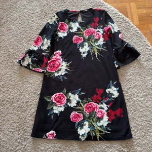 Svart blommig klänning från ONLY - Svart kortklänning från ONLY med stora blommor i rosa, vitt och rött. Klänningen har trekvartsärmar med volang och en snygg öppning med knapp i nacken. Tillverkad i ett mjukt, lätt material med innerfoder för extra komfort. Perfekt nu till hösten 🍁