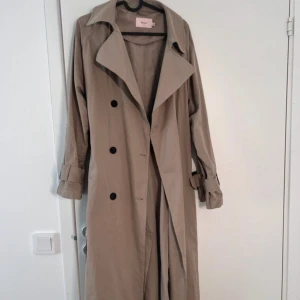 Beige trenchcoat från Nelly - Stilren beige trenchcoat från Nelly med klassisk krage, svarta knappar och justerbara ärmslut. Lång modell med snygg passform och bälte i midjan. Perfekt för lager-på-lager och en clean look.