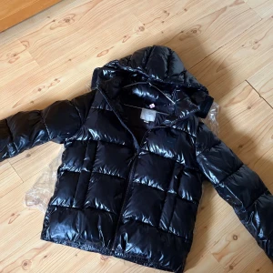Svart pufferjacka från Calvin Klein - Snygg svart pufferjacka från Calvin Klein med glansig finish och huva. Jackan har dragkedja framtill och breda, vadderade paneler som ger en cool och modern look. Perfekt för kalla dagar när du vill hålla stilen. Nypris 3000