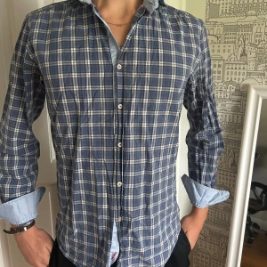 Blårutig skjorta från Stenströms - Säljer en blårutig skjorta från Stenströms med vita och grå rutor. Skjortan har klassisk krage, knappar framtill och ljusblå kontrasttyg på insidan av manschetterna. Broderad logga i rött på insidan. Perfekt för dig som gillar stilrena detaljer.