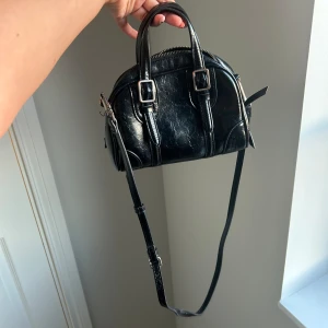 Svart liten handväska med axelrem - Snygg svart handväska i skinnimitation med rundad form och silverfärgade spännen framtill. Väskan har både handtag och justerbar axelrem, samt dragkedja upptill. Perfekt storlek för det viktigaste och passar till många olika outfits.