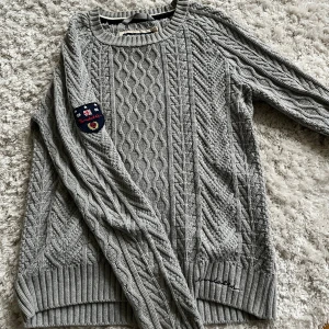 Grå stickad tröja från Bondelid - Säljer en grå stickad tröja från Bondelid med snyggt kabelstickat mönster och patch på ena ärmen. Tröjan har rund halsringning, ribbade muddar och är långärmad. Perfekt för kyliga dagar och riktigt skön att ha på sig.
