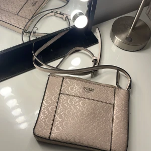 Beige axelväska från Guess med logomönster - Snygg beige axelväska från Guess med präglat logomönster över hela väskan. Väskan har justerbar axelrem, dragkedja upptill och insida med leopardmönstrat foder. Materialet är PVC och PU med polyesterfoder. Perfekt för dig som gillar stilrena detaljer och trendig design.