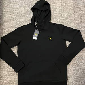 Svart hoodie från Lyle & Scott - Svart hoodie från Lyle & Scott med klassisk gul logga på bröstet. Tröjan har huva, känguruficka och långa ärmar. Tillverkad i mjuk bomullsmix som är perfekt för höst och vinter. Enkel och stilren design som funkar till allt.