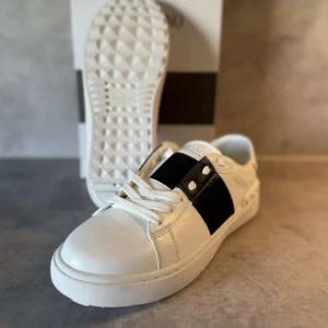 Valentino Garavani vita sneakers - Snygga vita sneakers från Valentino Garavani med bred svart rem över vristen och coola nitar på hälen. Skorna har rund tå, platt sula och klassisk snörning. Tillverkade i skinn för en lyxig känsla och stilren look. Perfekta för dig som vill sticka ut med designerstil.