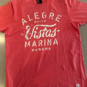 Rosa t-shirt Fisher and Bennet M - Snygg rosa t-shirt från Fisher and Bennet i storlek M. Tryck framtill med texten 'Alegre Vistas Marina Panama' i vitt. Klassisk passform med rund hals och korta ärmar. Perfekt för en chill och avslappnad stil.