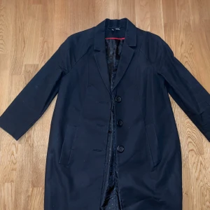 Svart kappa med knappar - Säljer en svart kappa med klassisk krage och tre stora knappar framtill. Jackan har raka ärmar och två fickor på sidorna. Perfekt för dig som gillar en stilren och enkel look. Fodrad insida för extra komfort.