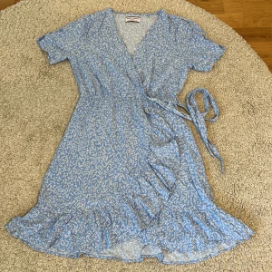 Blå omlottklänning Holly & Whyte - Söt blå kortklänning från Holly & Whyte med vitt blommigt mönster. Klänningen är med knyt i midjan och v-ringad hals. Det är en väldigt lätt klänning som är perfekt för sommaren 🌸☀️