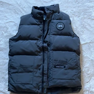 Grå  dunväst från Canada Goose - Svart dunväst från Canada Goose med hög krage och två stora fickor framtill. Västen har ett broderat märke på bröstet och stängs med både dragkedja och knappar. Yttermaterialet är polyester och bomull, fodret är nylon. Perfekt för kalla dagar.