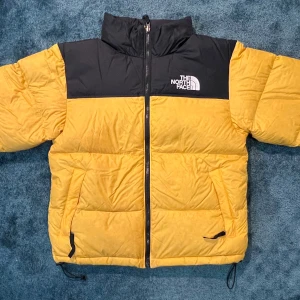 Gul och svart pufferjacka The North Face - Säljer en ikonisk pufferjacka från The North Face i gul och svart. Jackan har hög krage, dragkedja framtill, två fickor med dragkedja och justerbar nederkant. Perfekt för kalla dagar och riktigt snygg streetstil.