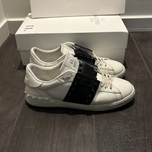 Valentino Garavani Rockstud sneakers - Snygga vita Valentino Garavani Rockstud sneakers med bred svart rem och nitar över vristen. Skorna har rund tå, snörning och är gjorda i skinn. Kommer med originalkartong och dustbag. Perfekta för dig som vill sticka ut med en lyxig och edgy stil. Använda i ungefär 2 veckor och är lite smuts på skosnörena och under skon. 