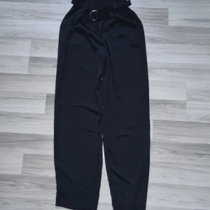 Svarta byxor med bälte från BikBok XS - Svarta byxor från BikBok i storlek XS med hög midja och papperspåse-linning. Byxorna har ett tygbälte med metallspänne och vida ben. Materialet är mjukt och lätt, perfekt för en chill och stilren look.