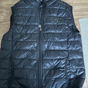Svart dunväst från EA7 Emporio Armani - Snygg svart dunväst från EA7 Emporio Armani med huva och dragkedja framtill. Västen har quiltad design och logga på bröstet. Perfekt för lager-på-lager och streetstyle. Materialet är glansigt och känns lätt men ändå värmande.