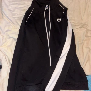 Svart tracksuit från Sergio Tacchini - Svart tracksuit från Sergio Tacchini med elastisk midja och diskret logga på benet. Byxorna är i mjukt material och har en klassisk sportig look, perfekta för chill eller träning. Matcha med tillhörande ziptröja för en komplett tracksuit.