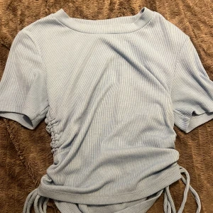 Ljusblå ribbad topp från Zara - Supersöt ljusblå ribbad topp från Zara med korta ärmar och snörning i sidorna som ger en snygg rynkad effekt. Toppen har en rund halsringning och är tillverkad i mjukt, stretchigt material. Perfekt för att skapa en chill och trendig look.