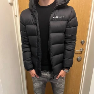 Svart pufferjacka Sail Racing - Säljer en svart pufferjacka från Sail Racing med huva och diskret logga på bröstet. Jackan har en klassisk quiltad look, dragkedja framtill och är perfekt för kyliga dagar. Passformen är normal och jackan är riktigt skön att bära.