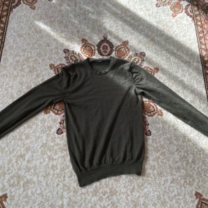 Merinotröja J.Lindeberg - Olivgrön stickad tröja från J.Lindeberg med rund halsringning och diskret broderad logga på bröstet. Tröjan har långa ärmar och ribbade muddar vid ärmslut och nederkant. 