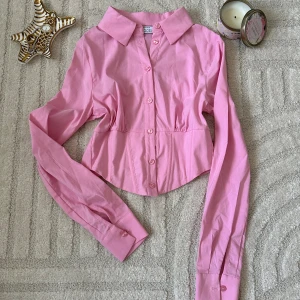 Rosa croppad skjorta med knappar - Snygg rosa croppad skjorta från Coco med klassisk krage och långa ärmar. Skjortan har markerad midja och knäppning framtill med matchande rosa knappar. Perfekt för dig som vill ha en trendig och feminin look.