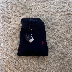 Mörkblå pikétröja från Polo Ralph Lauren - Mörkblå pikétröja från Polo Ralph Lauren med klassisk krage, tre vita knappar och röd broderad logga på bröstet. Tröjan är i bomull och har korta ärmar. Perfekt för en clean och stilren look.