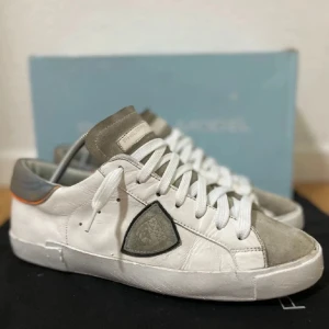 Vita Philippe Model skor - Snygga vita sneakers från Philippe Model med grå mockadetaljer på tå och plös. Skorna har en ikonisk sköldlogga på sidan, vita skosnören och diskret orange detalj vid hälen. Tillverkade i skinn och mocka för en lyxig känsla.