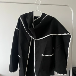 Svart jacka  med vit broderad kant - Svart kavaj med snygga vita broderade kanter längs krage, ärmar och fickor. Jackan har knappar framtill och två fickor. Den har en rak passform och lång ärm, perfekt för dig som vill sticka ut med en unik detalj.