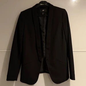 Svart kavaj från H&M, storlek 36 - Säljer en stilren svart kavaj från H&M i storlek 36. Kavajen har klassisk passform med slät framsida, smala slag och två fickor framtill. Aldrig använd