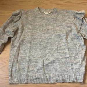 Grå stickad tröja med volang UNIQ Studio - Superfin ljusgrå stickad tröja från UNIQ Studio med volangdetaljer på axlarna. Tröjan har rund halsringning, ribbade muddar och långa ärmar. Perfekt för dig som vill ha en enkel men ändå trendig look.