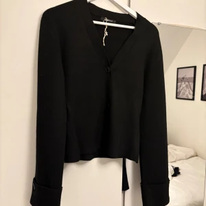 Viral gina tricot kofta - Stilren svart kofta från Weekday med v-ringad hals och knappstängning framtill. Långa ärmar med breda uppvik och dekorativa knappar vid ärmsluten. Perfekt till lager-på-lager och enkel att matcha med allt.