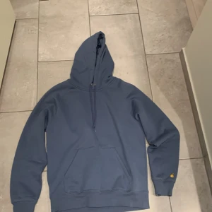 Blå Carhartt hoodie med huva - Säljer en blå hoodie från Carhartt med klassisk huva och snörning. Tröjan har en stor magficka och Carhartt-logga broderad i gult på ärmen. Tillverkad i mjuk bomull och har långa ärmar med ribbade muddar. Perfekt för chill dagar.