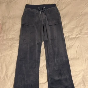 blå mjukisbyxor Juicy Couture - Blå mjukisbyxor från Juicy Couture i mjuk velour med bred resår och snörning i midjan. Byxorna har stora fickor fram och bak med knappdetaljer. Perfekta för en chill och avslappnad stil.