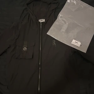 Svart vindjacka från Risky med huva - Svart vindjacka från Risky med huva och hel dragkedja framtill. Jackan har en cool bröstficka med spänne och Risky-logga på bröstet. Lätt och smidig med meshfoder på insidan, perfekt för blåsiga dagar.